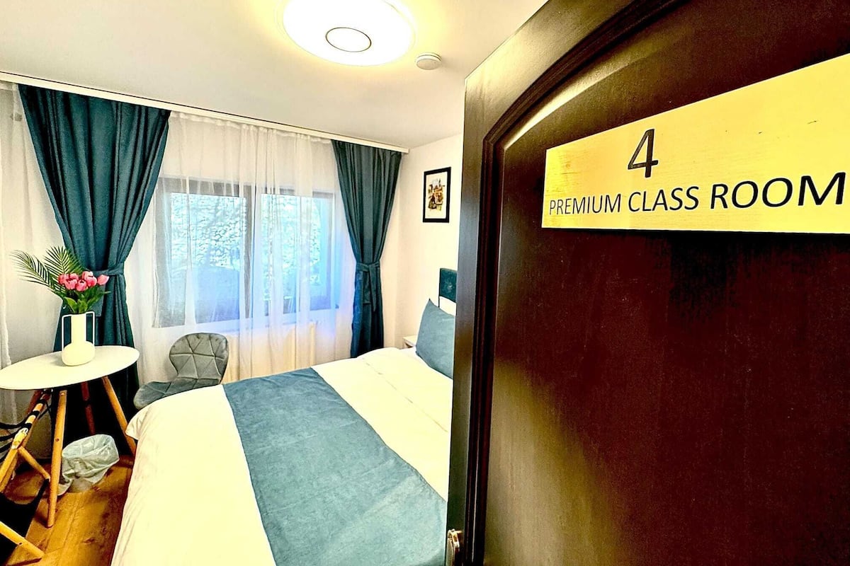 A room with a bed, a small table with a vase of flowers, and a window with curtains. A sign on the door reads "4 PREMIUM CLASS ROOM".