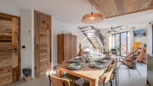 Un coin repas avec une table en bois dressée pour six personnes, entourée de chaises dotées de coussins à motifs. Un escalier en bois mène à un niveau supérieur, et une grande fenêtre offre une vue sur un paysage montagneux.