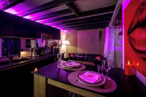 A dimly lit bar area with a table set for two, a sofa, and a decorative wall. - Love Room Perpignan • Jacuzzi, Tantra, Swing & Air Conditioning (Perpignan)