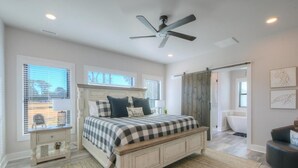 A bedroom with a bed, a ceiling fan, a window with blinds, a bedside table, and a bathroom visible through an open door. - The Bat Cave at OKI: Cozy 3BR retreat in Oak Island, near beaches and mini golf! (Oak Island)