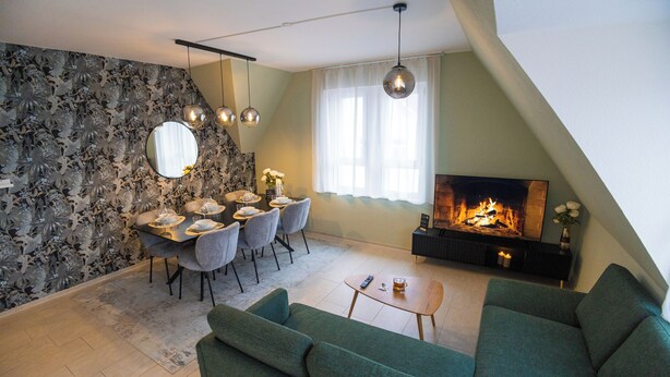 A modern living room with a dining area, green sofas, a fireplace, and a large mirror on the wall. - Duplex Apartment – Lake Constance – Self-Check-In – Parking – 4-Keys Apartments (Rorschach)