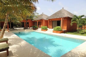 A swimming pool area with two thatched-roof structures, palm trees, and a bench. - Villa SUNUGAL (Ngaparou)