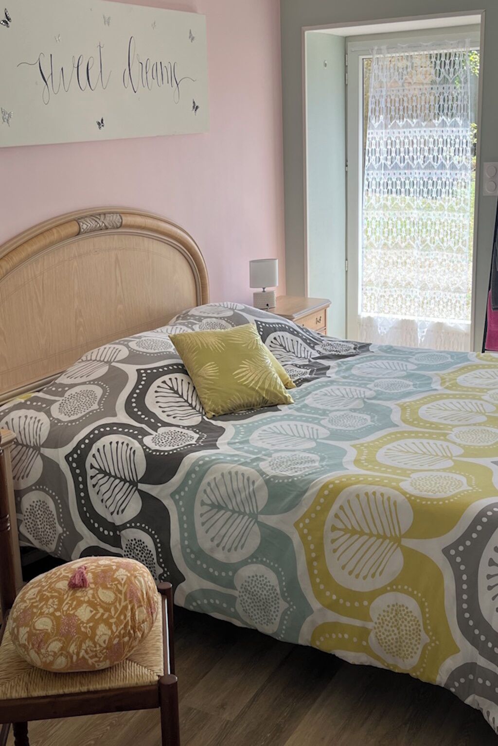 Un dormitorio con cama, una silla, una mesita de noche, una lámpara y un ventanal con cortina de encaje.