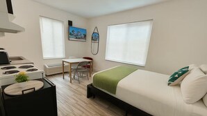 Une petite pièce comprenant un lit, un petit bureau, une chaise, une cuisinière et deux fenêtres équipées de stores.