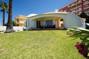 A modern villa with a curved roof, a patio with outdoor furniture, and a well-maintained lawn. - Beachfront location - Ideal family home - Pool & garden (Armação de Pêra)