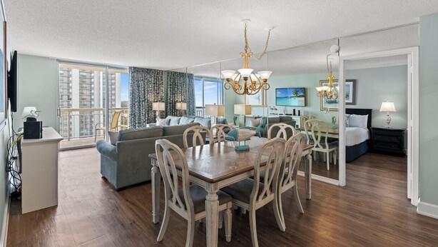 A spacious living room with a dining area, a large window with a view of high-rise buildings, and a chandelier. - S Hampton 1406 2 Br Oceanview in Kingston, Pool (Myrtle Beach)