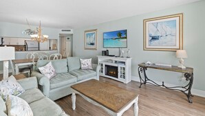 A living room with a sofa, coffee table, and fireplace. There are framed pictures on the wall and a dining area in the background. - S Hampton 1406 3 Br Oceanview in Kingston, Pool (Myrtle Beach)