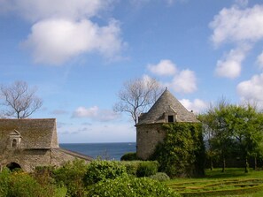 A stone building with a conical roof and a smaller adjacent structure, surrounded by greenery and a clear sky. - Mill for 2, facing the sea, 300m to the beach and great walks. Next to a castle. (La Hague)
