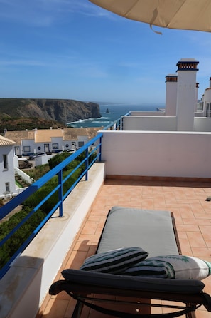 A balcony with a view of the ocean, a lounge chair, and a blue railing.