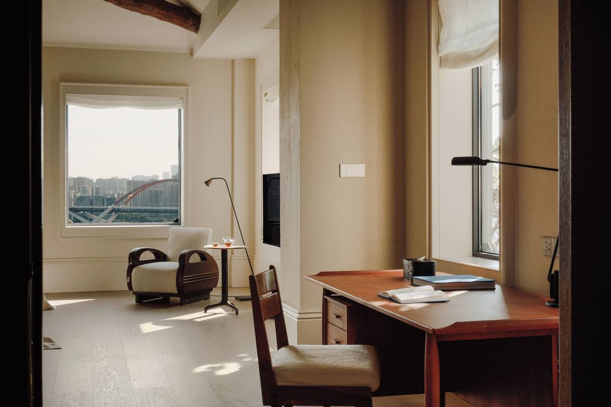 Een kamer met een bureau, twee stoelen, een raam met uitzicht op een stadsgezicht en een lamp.