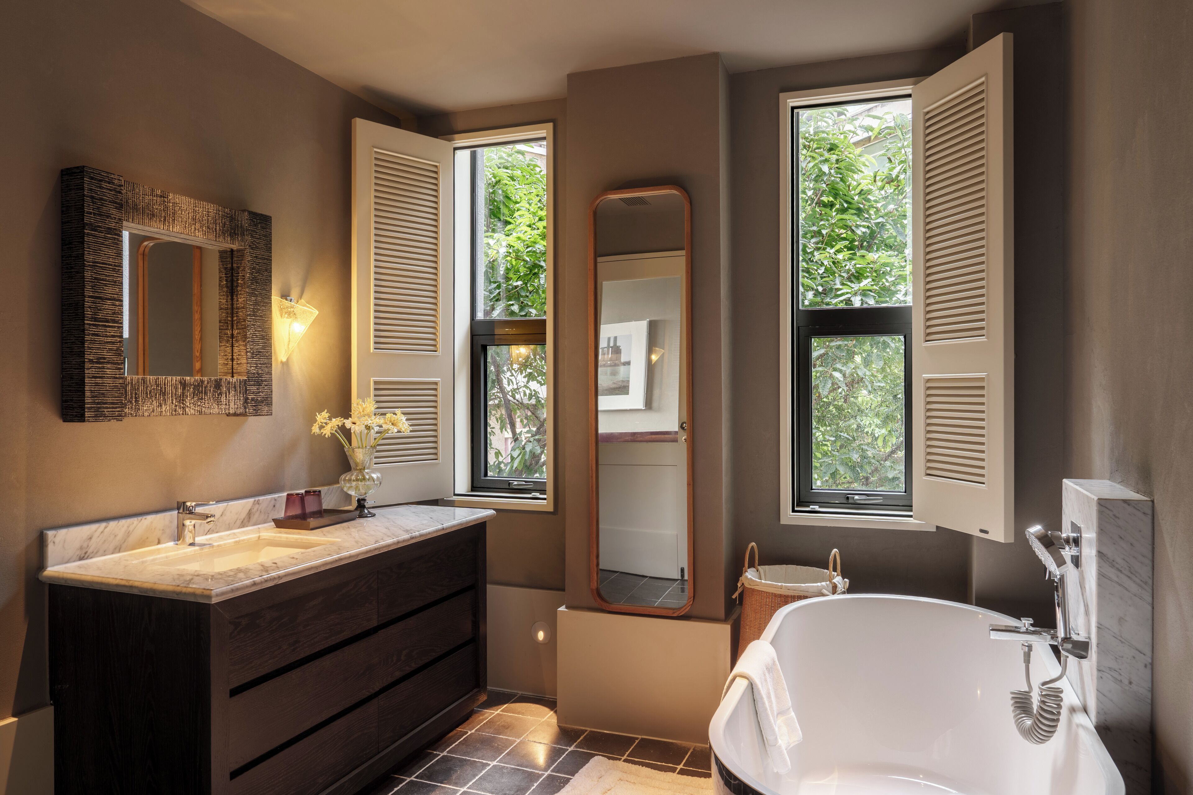 A modern bathroom with a marble sink, dark wooden vanity, a freestanding bathtub, and large windows with shutters.
