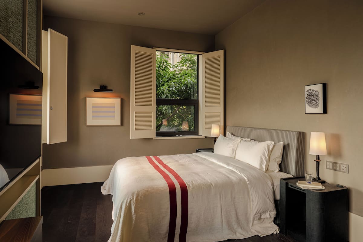 Een verzorgde slaapkamer met een groot bed, een raam met geopende luiken, een nachtkastje met een lamp en een lijst met een afbeelding aan de muur.