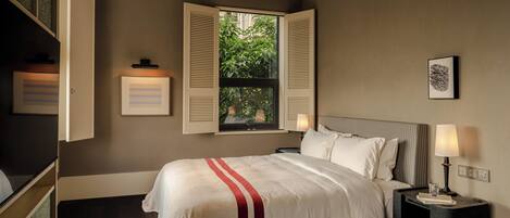 A neatly arranged bedroom with a large bed, a window with open shutters, a bedside table with a lamp, and a framed picture on the wall.