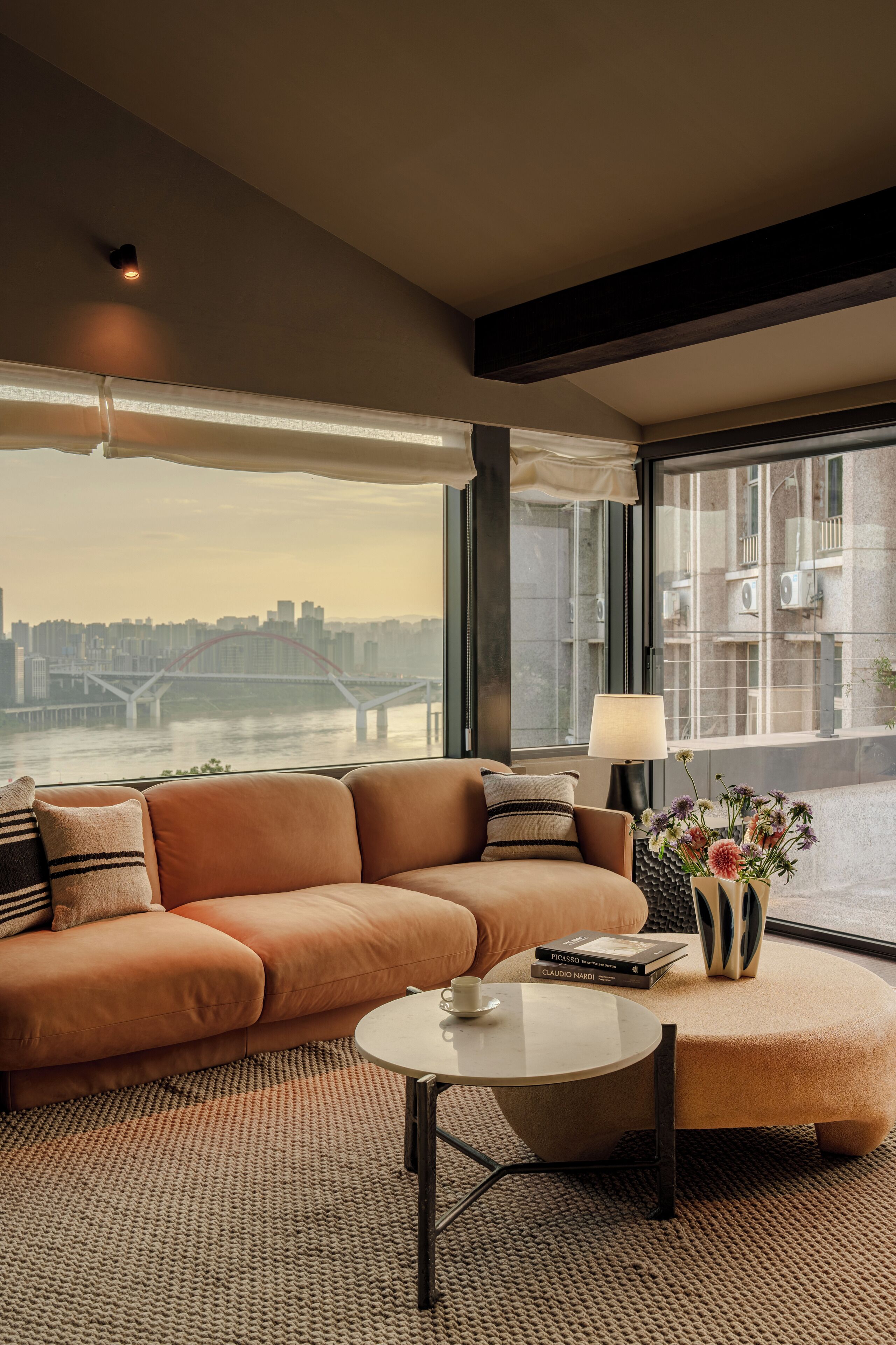 Un salon moderne avec un canapé, une table basse ronde et une lampe. Une fenêtre offre une vue sur le paysage urbain et un pont.