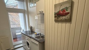 A compact kitchen with a washing machine, a sink, and a window overlooking a balcony.