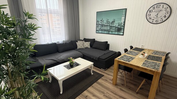 A modern living room with a sectional sofa, a coffee table, a dining table with chairs, a clock on the wall, and a cityscape painting.