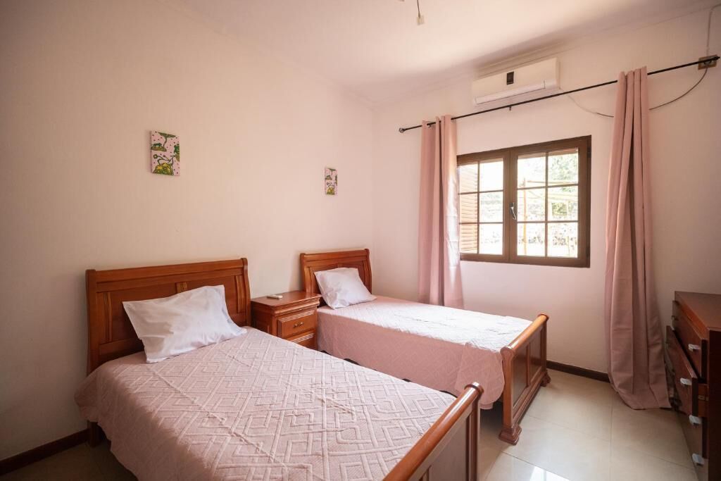 A room with two beds, a window with curtains, a wooden dresser, and a wall-mounted air conditioning unit.