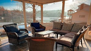 En overdækket terrasse med fletningsmøbler, et glasbord og udsigt til en sø og bygninger.