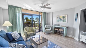 A living room with a blue sofa, a coffee table, a chair, a small table with stools, a ceiling fan, and a view of palm trees through large windows. - Southwind 204 1 Bedroom Condo (Myrtle Beach)