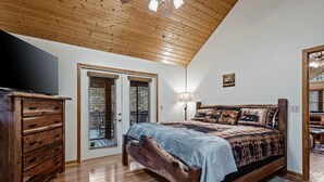 Une chambre avec un plafond en bois, un lit recouvert d’une couvre-lit à motifs, une commode en bois, un téléviseur et une fenêtre avec des stores.