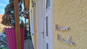 Een gebouwde ingang met een deur, een wandbord waarop 'Maison Marabu' staat, en een roze prullenbak.