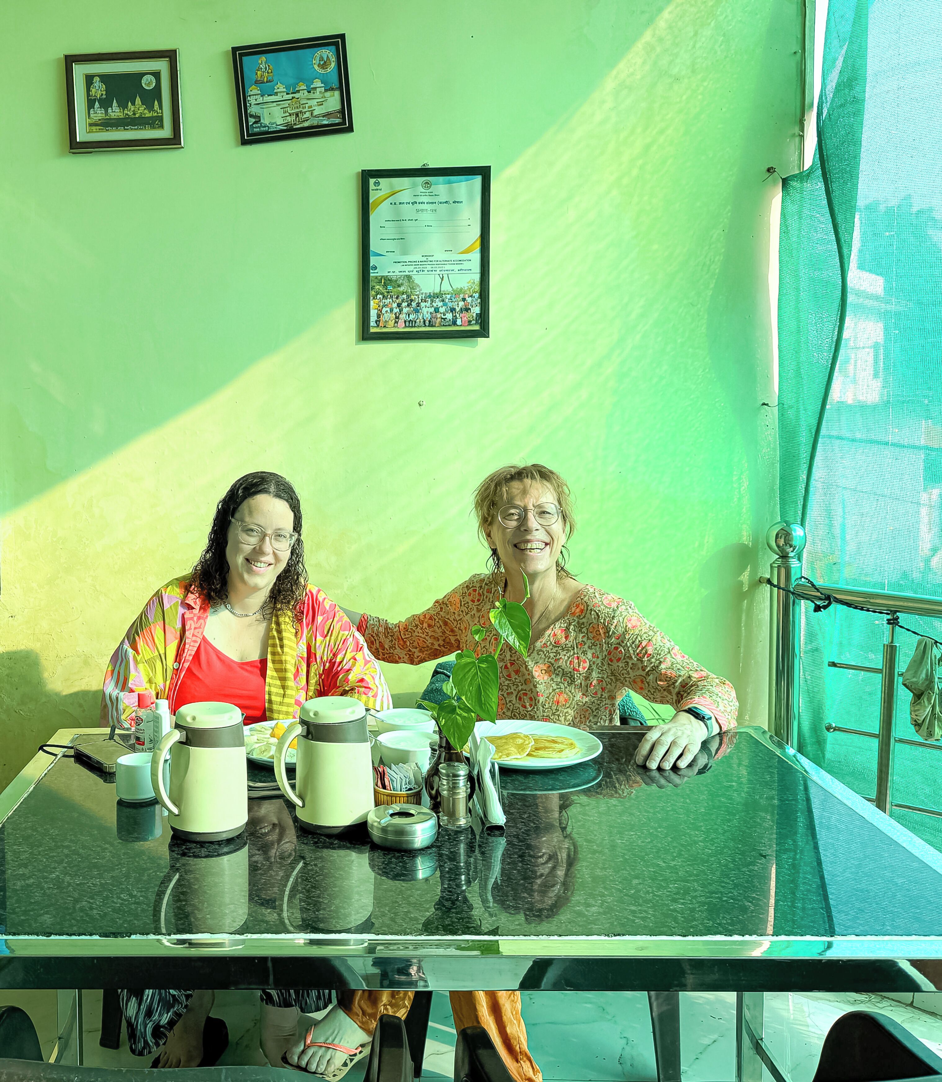 Two people seated at a table with a green wall behind them, a framed certificate, and a window with a mesh screen.