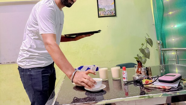 A person serving a beverage at a counter with a tray. The counter has a glass, a bottle, and a small bowl. There are framed certificates on the wall behind. - Orchha best homestay (Orchha)