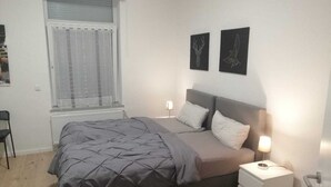 Une chambre à coucher comprenant un lit, une table de chevet, une fenêtre avec des rideaux et deux tableaux accrochés au mur.
