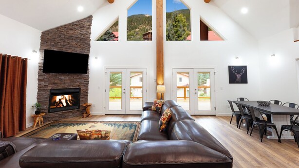 A spacious living room with a fireplace, leather sofa, dining table, and a high ceiling with exposed beams. - Almost Heaven - Newly Built! - In Town - On the River - WiFi - Grill - Mountain (Red River)