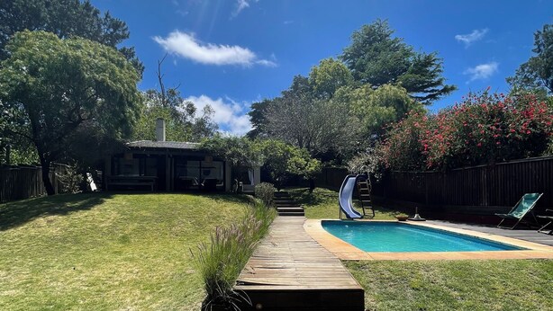 A backyard with a wooden deck leading to a swimming pool, surrounded by trees and a fence.
