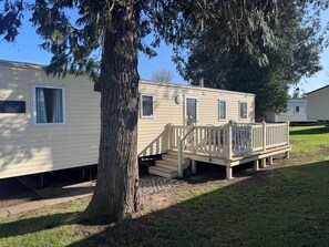 A mobile home with a deck, a tree in front, and a clear blue sky. - Cozy Caravan at Tower Lawn, Haggerston Castle Perfect Family Getaway (Berwick-upon-Tweed)
