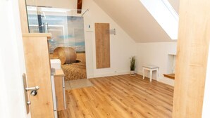 Una habitación con suelo de madera, una puerta de madera, una pequeña mesa blanca, una planta en maceta y un gran ventanal con vistas a pacas de heno.