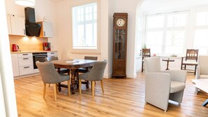 A spacious kitchen with wooden flooring, a dining table with chairs, a large grandfather clock, and a view of the outdoors through large windows. - Juliane | Charming Apartment with a Bay Window | North Sea (Norden)
