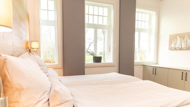 A bedroom with a bed, two windows, a nightstand, and a wall-mounted lamp. - Juliane | Charming Apartment with a Bay Window | North Sea (Norden)