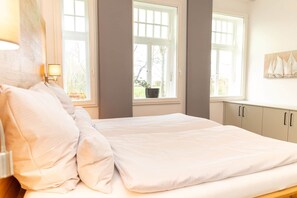 A bedroom with a bed, two windows, a nightstand, and a wall-mounted lamp. - Juliane | Charming apartment with a picture window | North Sea (Norden)