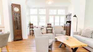 A spacious living room with a wooden floor, a large clock, and a view of the outdoors through large windows. - Juliane | Charming Apartment with a Bay Window | North Sea (Norden)