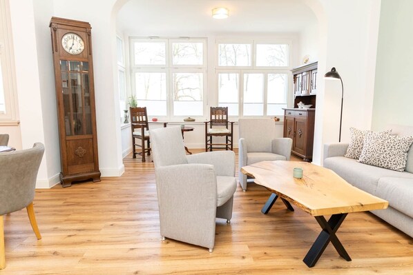 A spacious living room with a wooden floor, a large clock, and a view of the outdoors through large windows. - Juliane | Charming apartment with a picture window | North Sea (Norden)