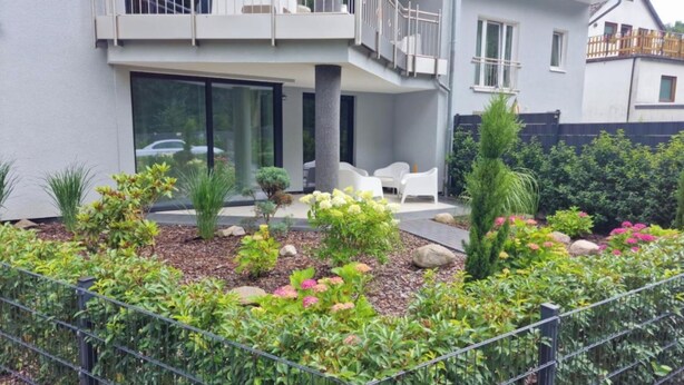 Ett modernt hus i två våningar med en balkong, en uteplats med utemöbler och en välskött trädgård med olika växter och blommor.