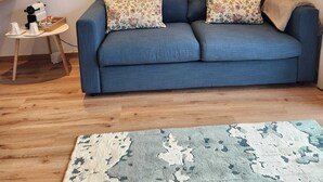 A living room with a blue sofa, two patterned cushions, a small side table with a coffee maker, and a rug. There are three framed artworks on the wall above the sofa.