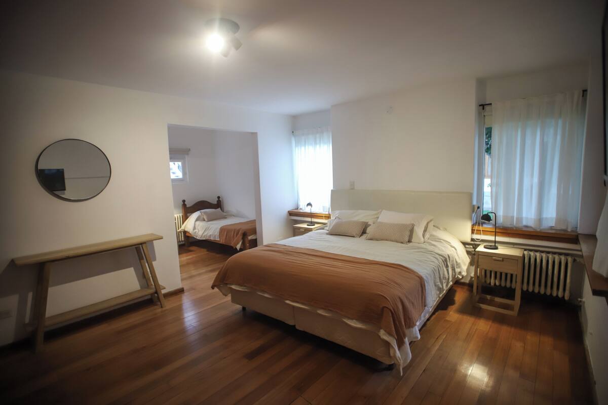A bedroom with two beds, a bedside table with a lamp, a round mirror, and a window with curtains.