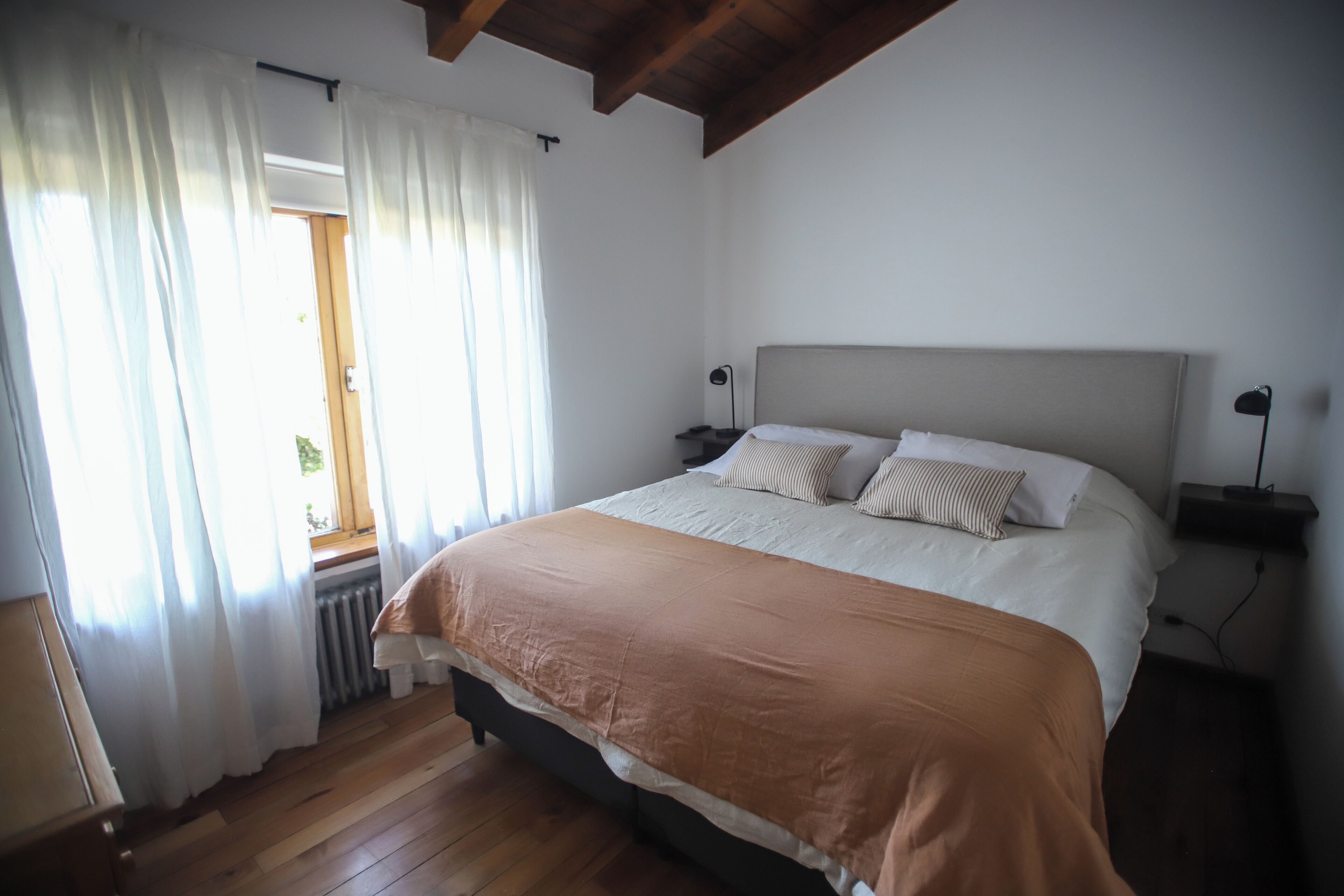 Een slaapkamer met een groot bed, witte gordijnen, een houten vloer en een raam met uitzicht.