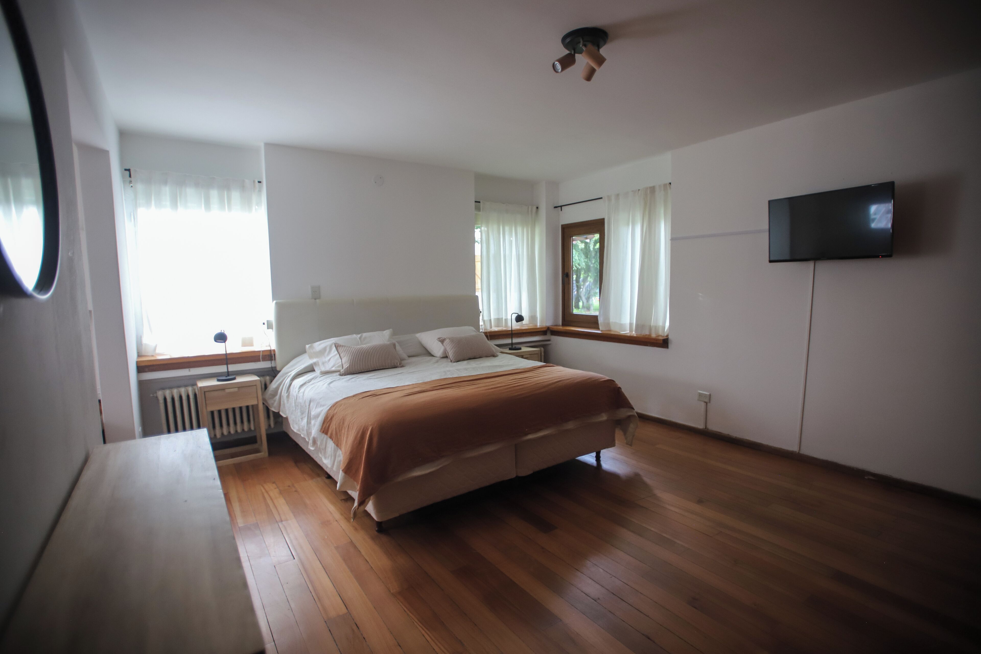 Un dormitorio con suelo de madera, una cama con manta marrón, una mesita de noche, un televisor montado en la pared y una ventana con cortinas.