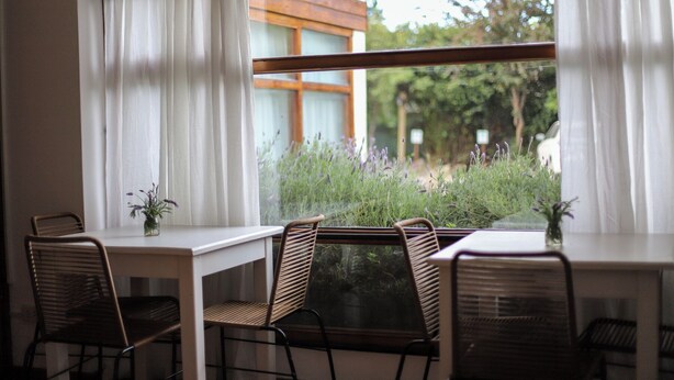 A dining area with a table and chairs, a window with white curtains, and a view of greenery and a building outside. - Hostería & Apart Hotel Carelhue (San Carlos de Bariloche)