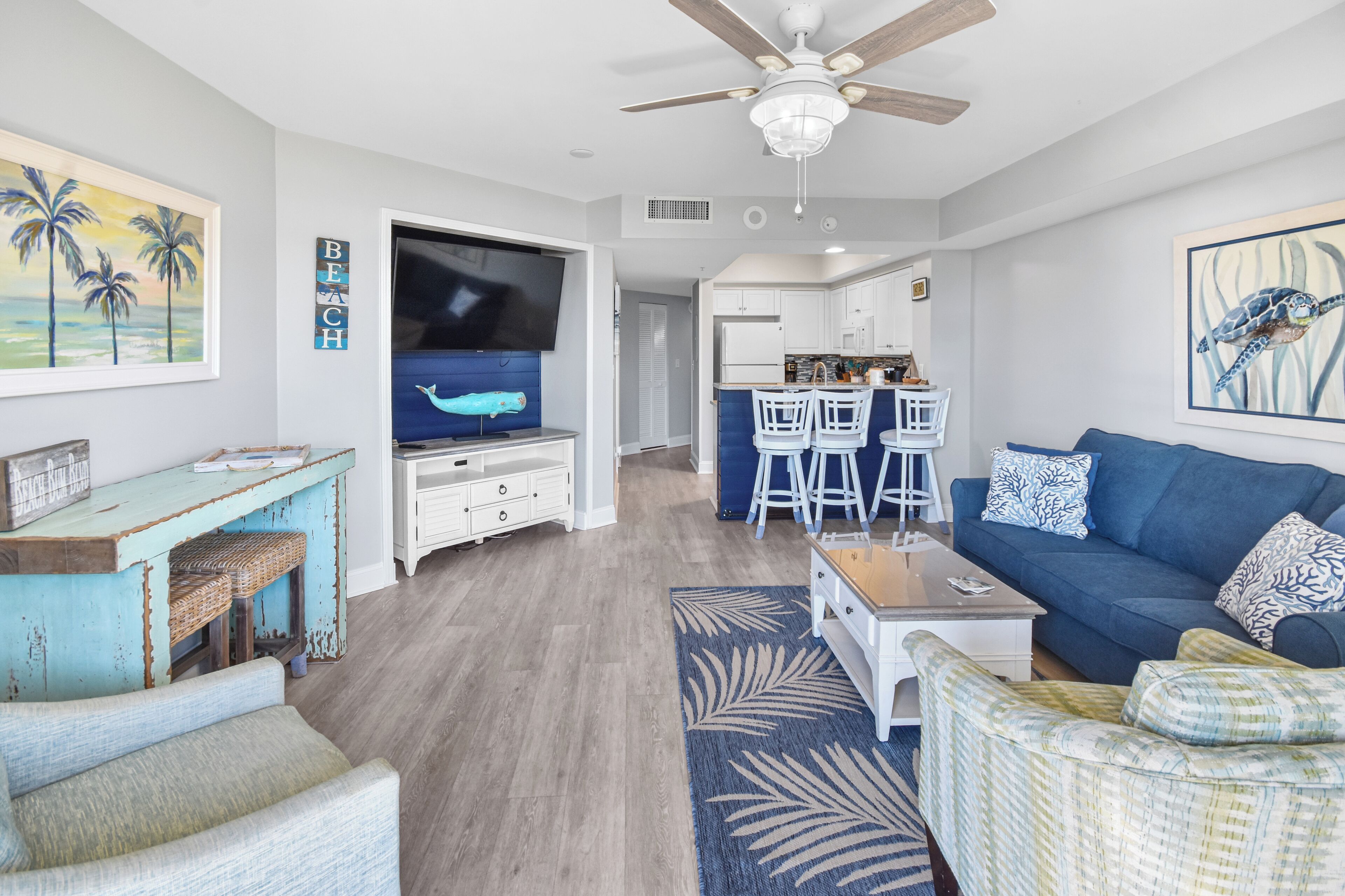 Una sala moderna con un sofá azul, una mesa de centro y un televisor. Hay una zona de cocina con armarios blancos y una mesa de comedor con taburetes de bar.