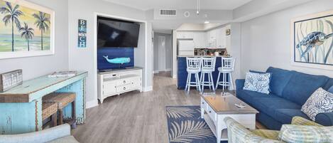 Una sala moderna con un sofá azul, una mesa de centro y un televisor. Hay una zona de cocina con armarios blancos y una mesa de comedor con taburetes de bar.