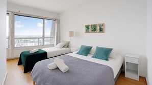 Een hotelkamer met twee bedden, een groot raam met uitzicht over de stad, een nachtkastje en drie ingelijste foto's aan de muur.