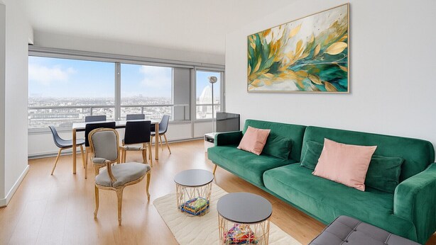 Een ruime woonkamer met een groene bank, een eettafel met stoelen en een groot raam met uitzicht op de stad.