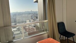 Een kamer met een groot raam, een stoel en een bed met een oranje deken.