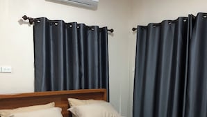 Una cama bien tendida con cabecera de madera, aire acondicionado de pared y cortinas oscuras a cada lado.