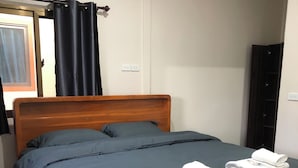 Una cama individual con estructura de madera y ropa de cama oscura, dos toallas sobre la cama, una ventana con cortinas negras y un aire acondicionado instalado en la pared.